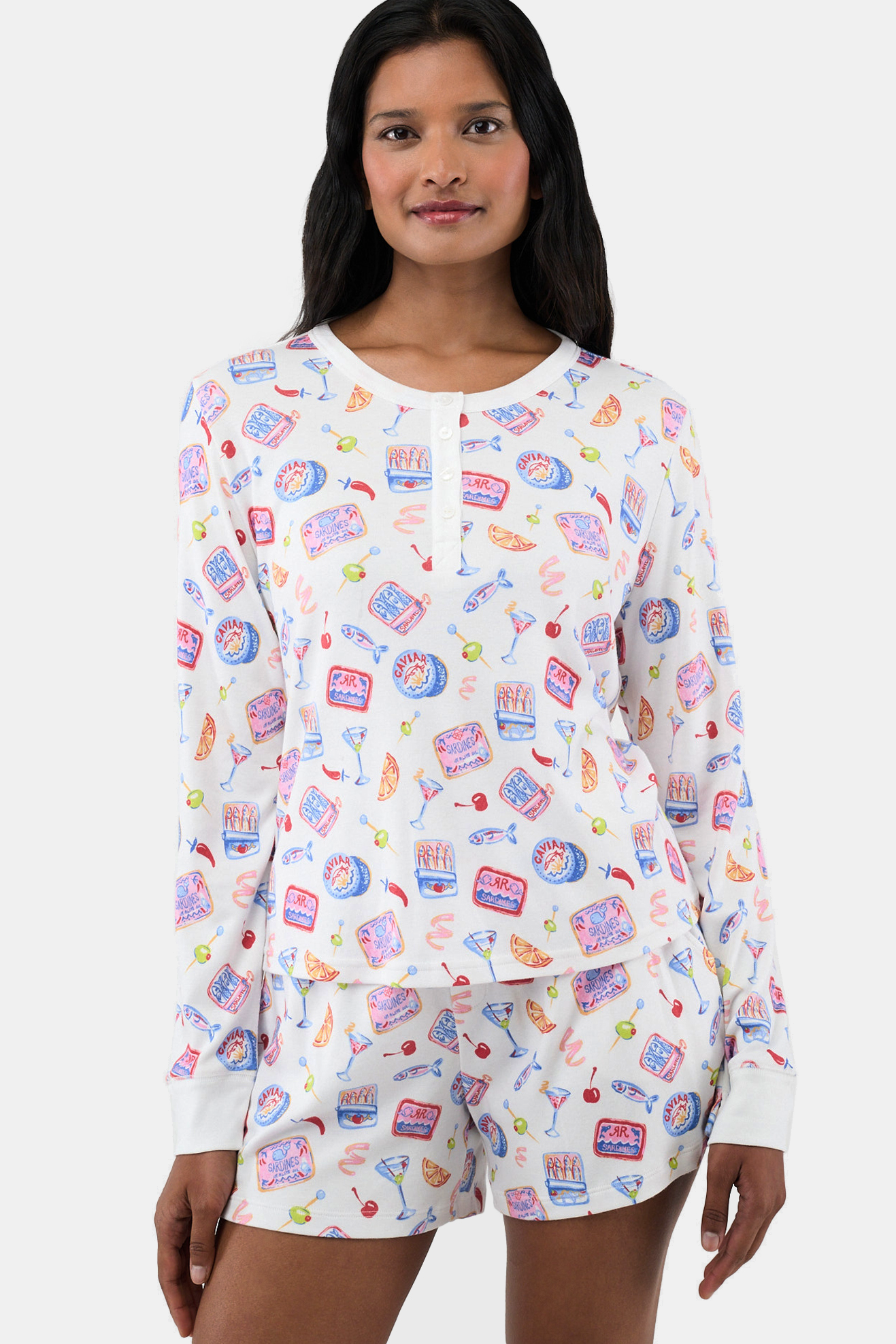 A woman smiles, wearing the Tinned or Twist Ashley Top—a Pima-modal henley pajama set with matching shorts, adorned with colorful caviar and drink motifs on crisp white fabric.