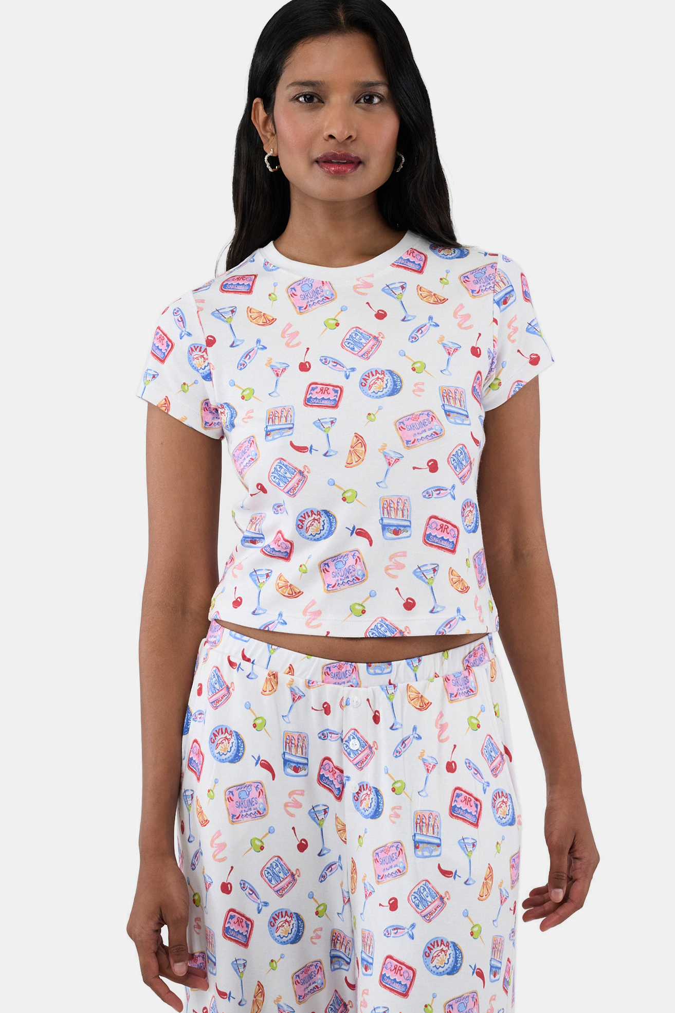 A woman with long dark hair wears a matching Brooklyn Boxer set and the Tinned or Twist Baby Tee, both adorned with colorful foodie prints—cocktails, canned drinks, lemon slices, and shrimp—while standing against a plain white background.