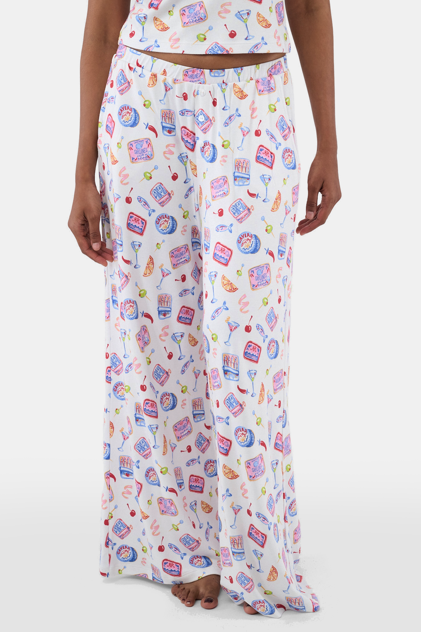 A person wears the Tinned or Twist Boden Pant—ultra-soft, loose white pajama pants featuring a colorful snack, drink, and food print—standing against a white background.