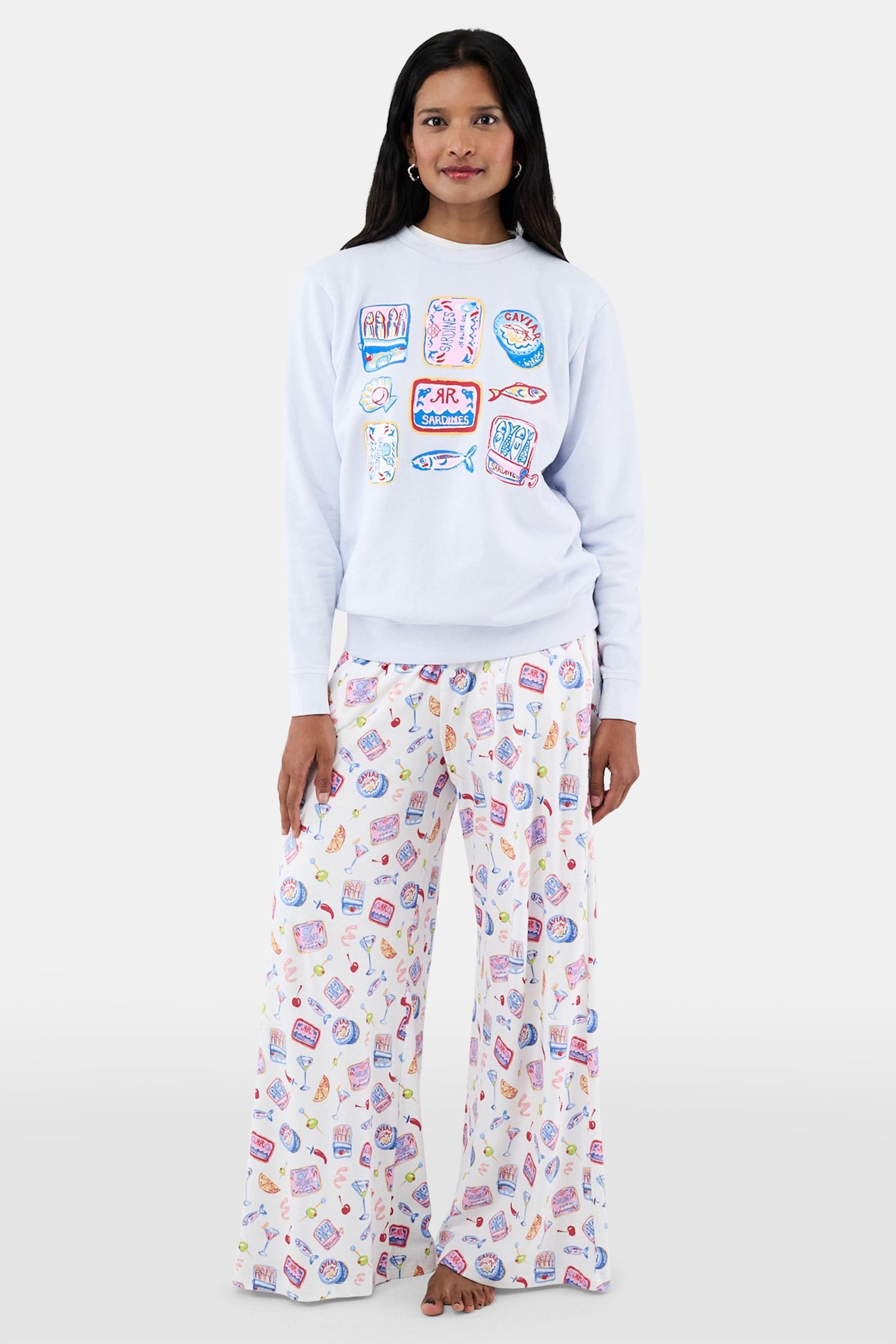 A woman with long dark hair wears the Roller Rabbit Tinned or Twist Crewneck, a light blue cotton fleece pullover with colorful sardine can and fish illustrations, paired with matching white pants featuring food-themed patterns against a plain white background.