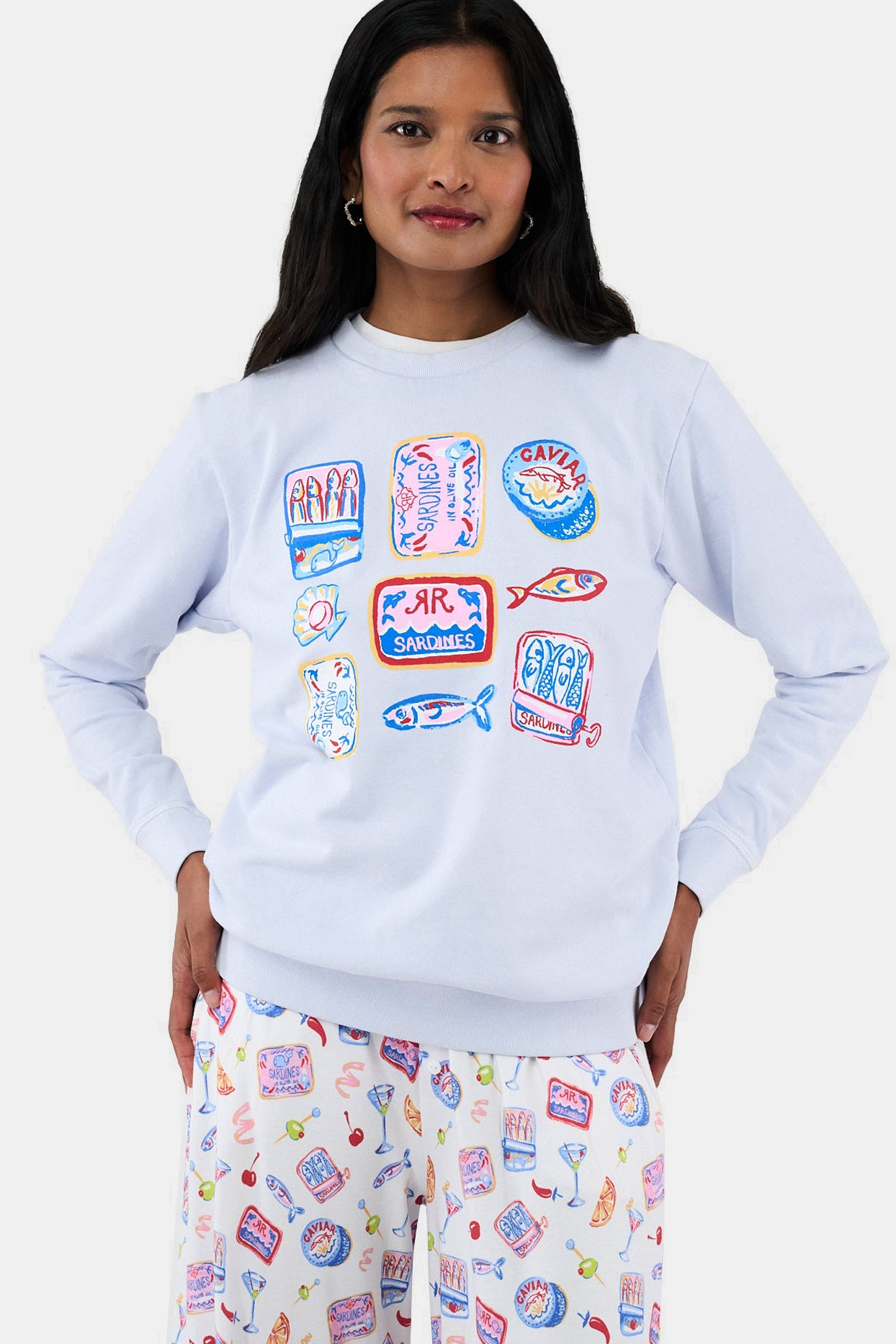 A woman with long dark hair wears the Roller Rabbit Tinned or Twist Crewneck, a light blue cotton fleece pullover with colorful sardine can and fish illustrations, paired with matching white pants featuring food-themed patterns against a plain white background.