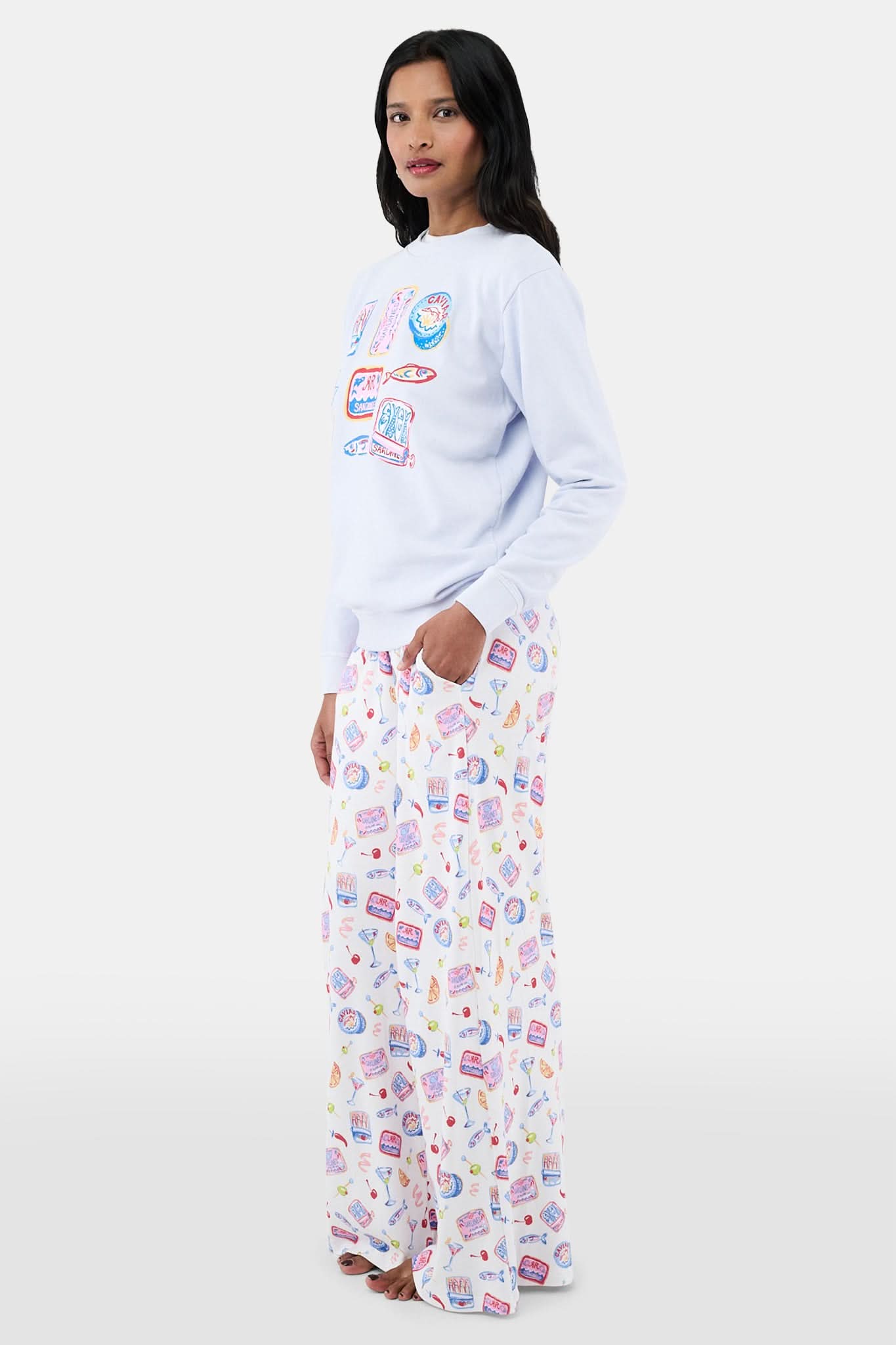 A woman with long dark hair wears the Roller Rabbit Tinned or Twist Crewneck, a light blue cotton fleece pullover with colorful sardine can and fish illustrations, paired with matching white pants featuring food-themed patterns against a plain white background.
