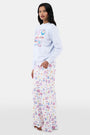 A woman with long dark hair wears the Roller Rabbit Tinned or Twist Crewneck, a light blue cotton fleece pullover with colorful sardine can and fish illustrations, paired with matching white pants featuring food-themed patterns against a plain white background.