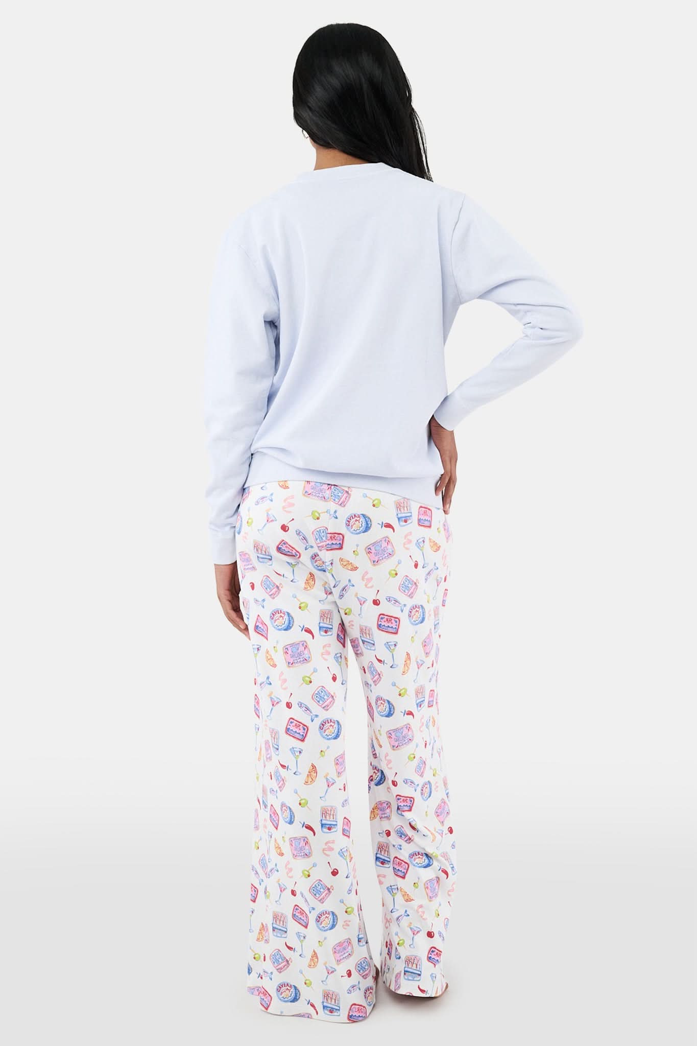 A woman with long dark hair wears the Roller Rabbit Tinned or Twist Crewneck, a light blue cotton fleece pullover with colorful sardine can and fish illustrations, paired with matching white pants featuring food-themed patterns against a plain white background.