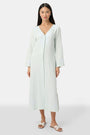 A woman faces forward wearing the Roller Rabbit Vanna Embroidery Rachel Caftan—a long, light green linen dress with subtle white embroidery and long sleeves—paired with white sandals against a plain light gray background.