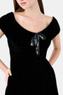 A woman faces forward in the Roller Rabbit Velvet Tilly Dress, a short-sleeve black velvet mini with a floral bow at the neckline, paired with black platform sandals, set against a plain light background.