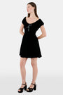 A woman faces forward in the Roller Rabbit Velvet Tilly Dress, a short-sleeve black velvet mini with a floral bow at the neckline, paired with black platform sandals, set against a plain light background.
