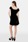 A woman faces forward in the Roller Rabbit Velvet Tilly Dress, a short-sleeve black velvet mini with a floral bow at the neckline, paired with black platform sandals, set against a plain light background.