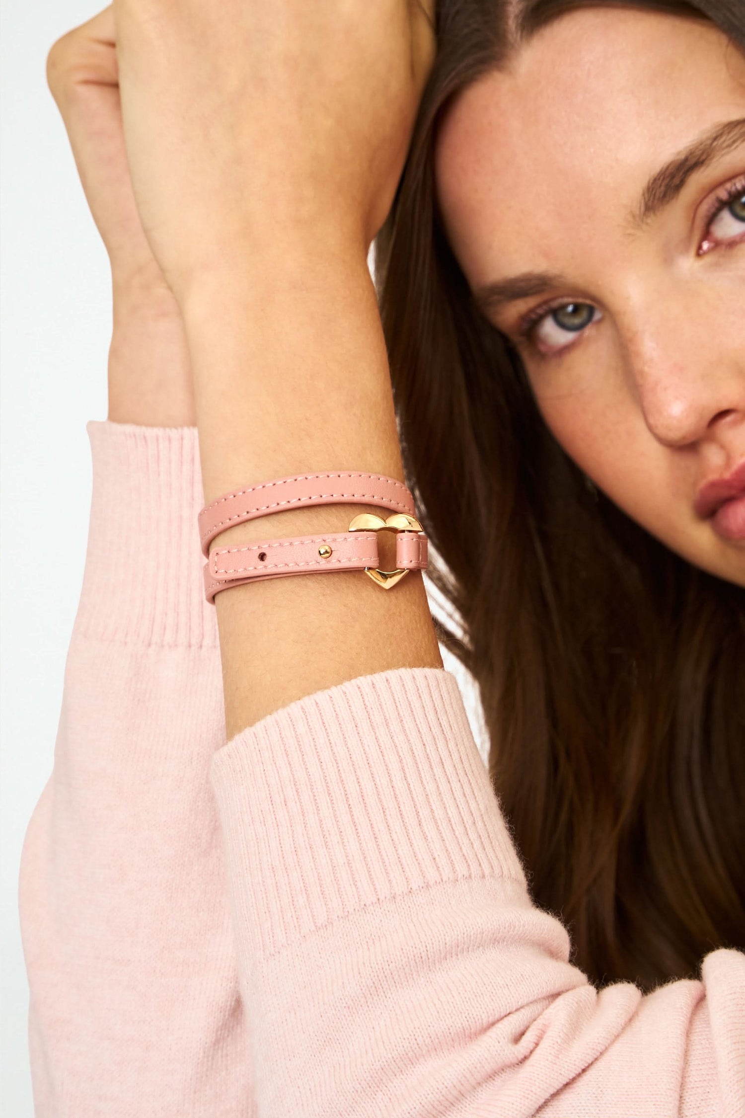 The Heather Love Bracelet by Roller Rabbit features pink leather with white stitching and an imitation gold heart-shaped clasp, shown coiled on a plain light background.