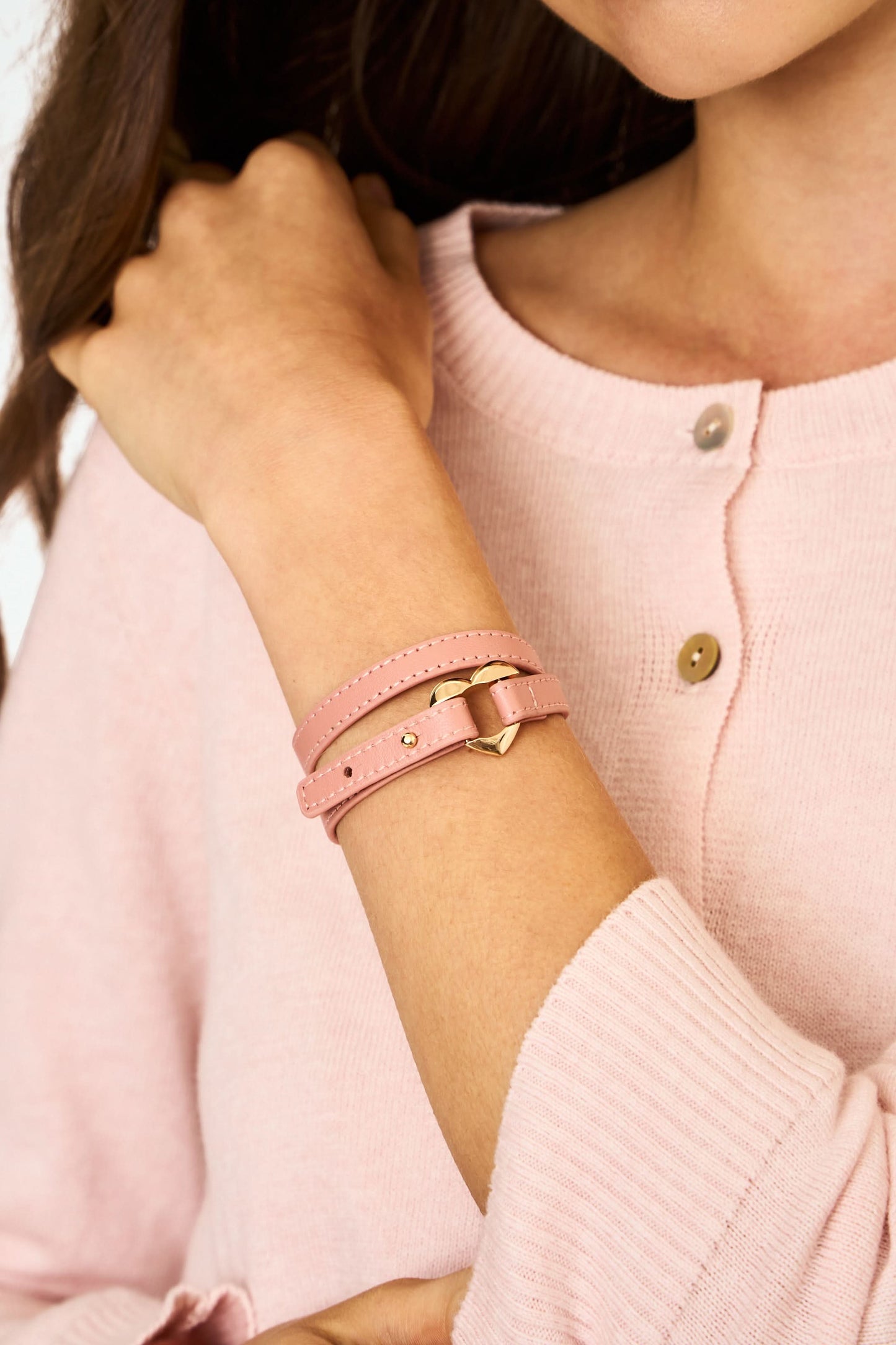 The Heather Love Bracelet by Roller Rabbit features pink leather with white stitching and an imitation gold heart-shaped clasp, shown coiled on a plain light background.