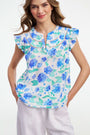 A smiling person wears the Roller Rabbit Hydrangea Emerson Top featuring blue hydrangeas and flutter sleeves, paired with white pants against a light, plain background.