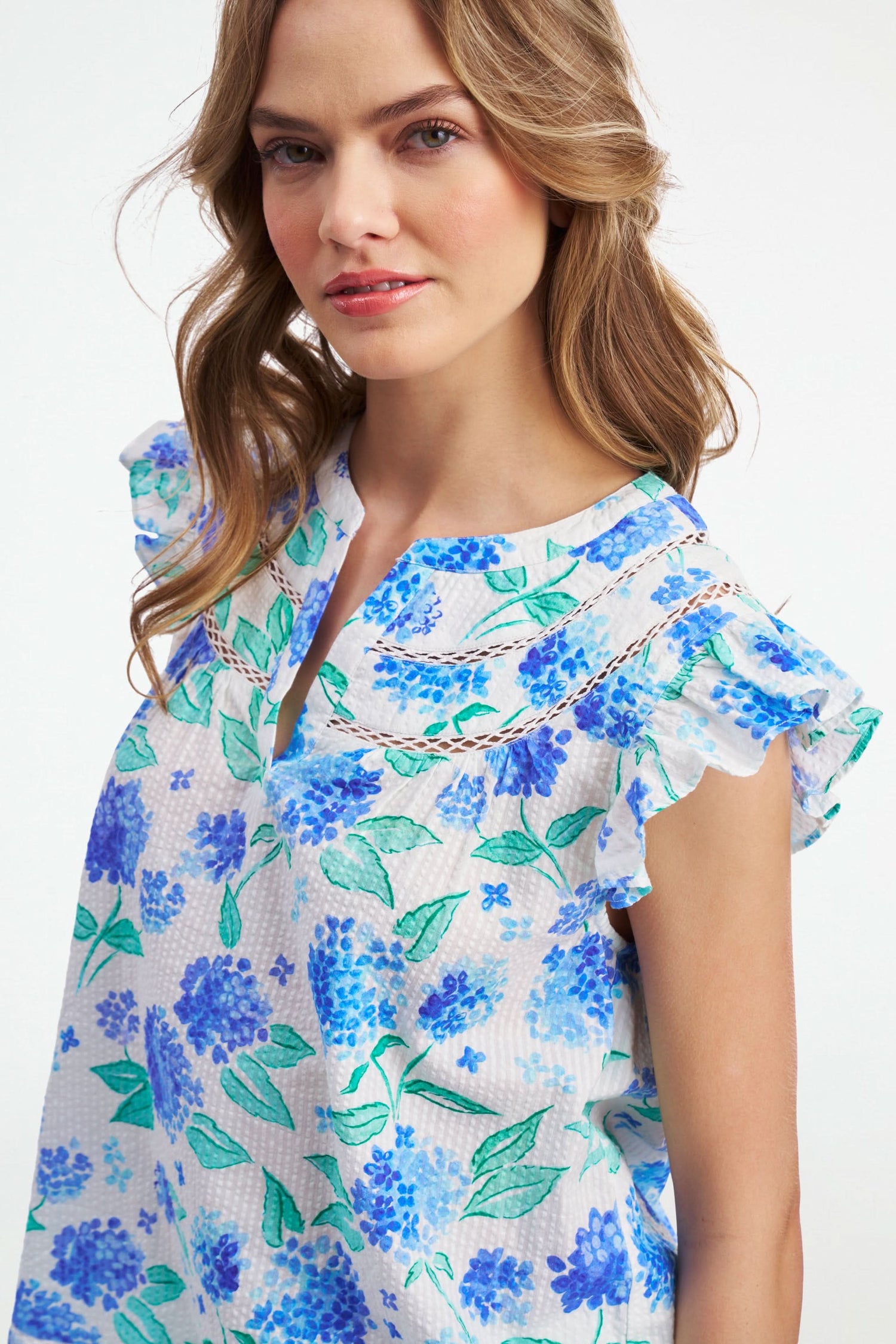 A smiling person wears the Roller Rabbit Hydrangea Emerson Top featuring blue hydrangeas and flutter sleeves, paired with white pants against a light, plain background.