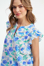 A smiling person wears the Roller Rabbit Hydrangea Emerson Top featuring blue hydrangeas and flutter sleeves, paired with white pants against a light, plain background.
