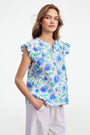 A smiling person wears the Roller Rabbit Hydrangea Emerson Top featuring blue hydrangeas and flutter sleeves, paired with white pants against a light, plain background.