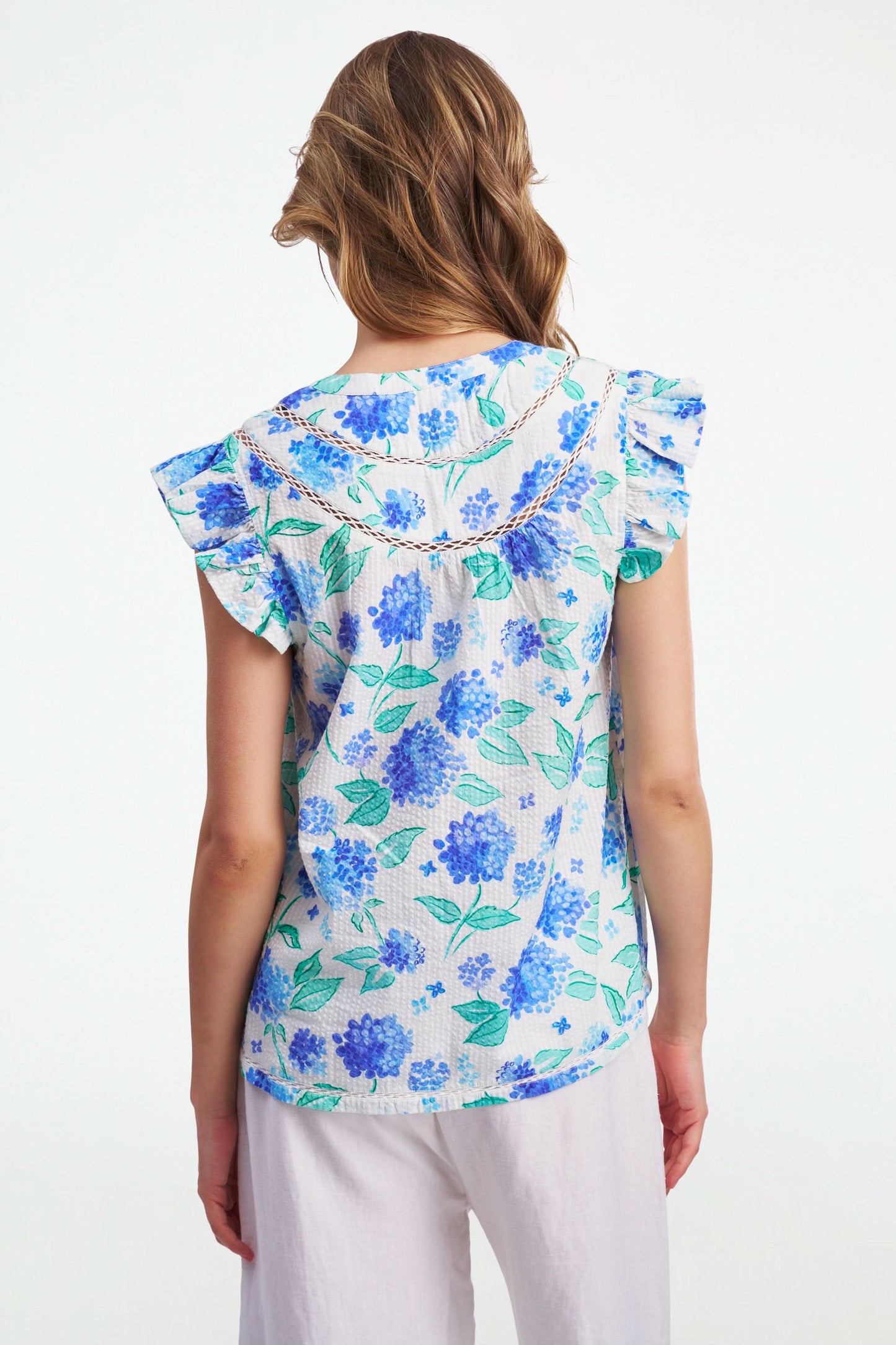 A smiling person wears the Roller Rabbit Hydrangea Emerson Top featuring blue hydrangeas and flutter sleeves, paired with white pants against a light, plain background.