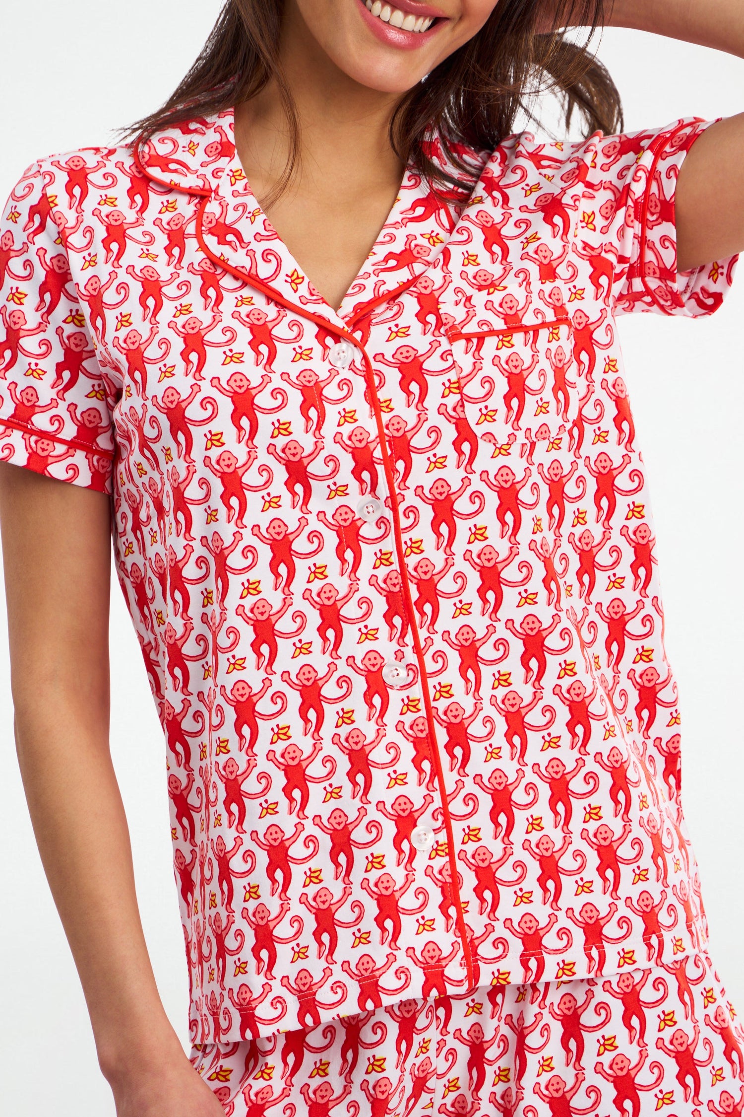 A woman wearing Monkey Polo Pajamas—short-sleeved, polo-style top and bottoms with a playful red monkey print on white—smiles while touching her hair.