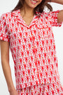 A woman wearing Monkey Polo Pajamas—short-sleeved, polo-style top and bottoms with a playful red monkey print on white—smiles while touching her hair.