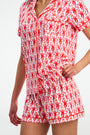 Someone wears the Monkey Polo Pajamas—a short-sleeve button-up shirt and shorts set made from soft pima cotton, featuring a red and pink monkey print on white. The image is cropped to show from the shoulders down.