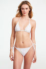 A person wears the Roller Rabbit Palm Dreams Serena Bikini Bottom, showcasing pastel palm trees and side ties, with one hand on their hip against a plain background.