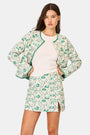 A person wears the Roller Rabbit Woodland Toile Quilted Azure Mini Skirt, featuring green floral patterns, green trim, a side slit, and paired with a matching white jacket—an ideal fall fashion ensemble.