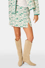 A person wears the Roller Rabbit Woodland Toile Quilted Azure Mini Skirt, featuring green floral patterns, green trim, a side slit, and paired with a matching white jacket—an ideal fall fashion ensemble.