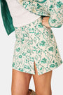 A person wears the Roller Rabbit Woodland Toile Quilted Azure Mini Skirt, featuring green floral patterns, green trim, a side slit, and paired with a matching white jacket—an ideal fall fashion ensemble.