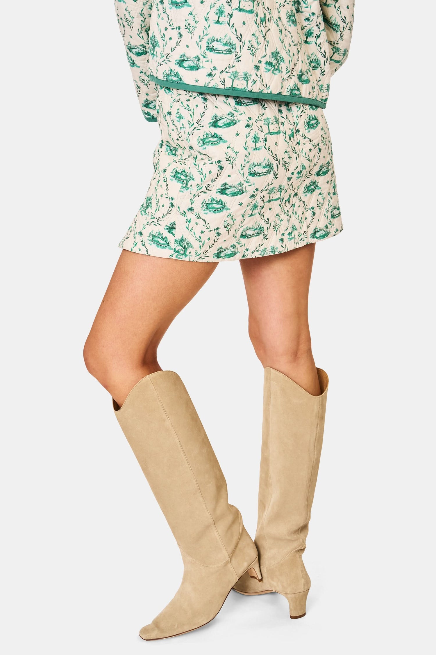 A person wears the Roller Rabbit Woodland Toile Quilted Azure Mini Skirt, featuring green floral patterns, green trim, a side slit, and paired with a matching white jacket—an ideal fall fashion ensemble.