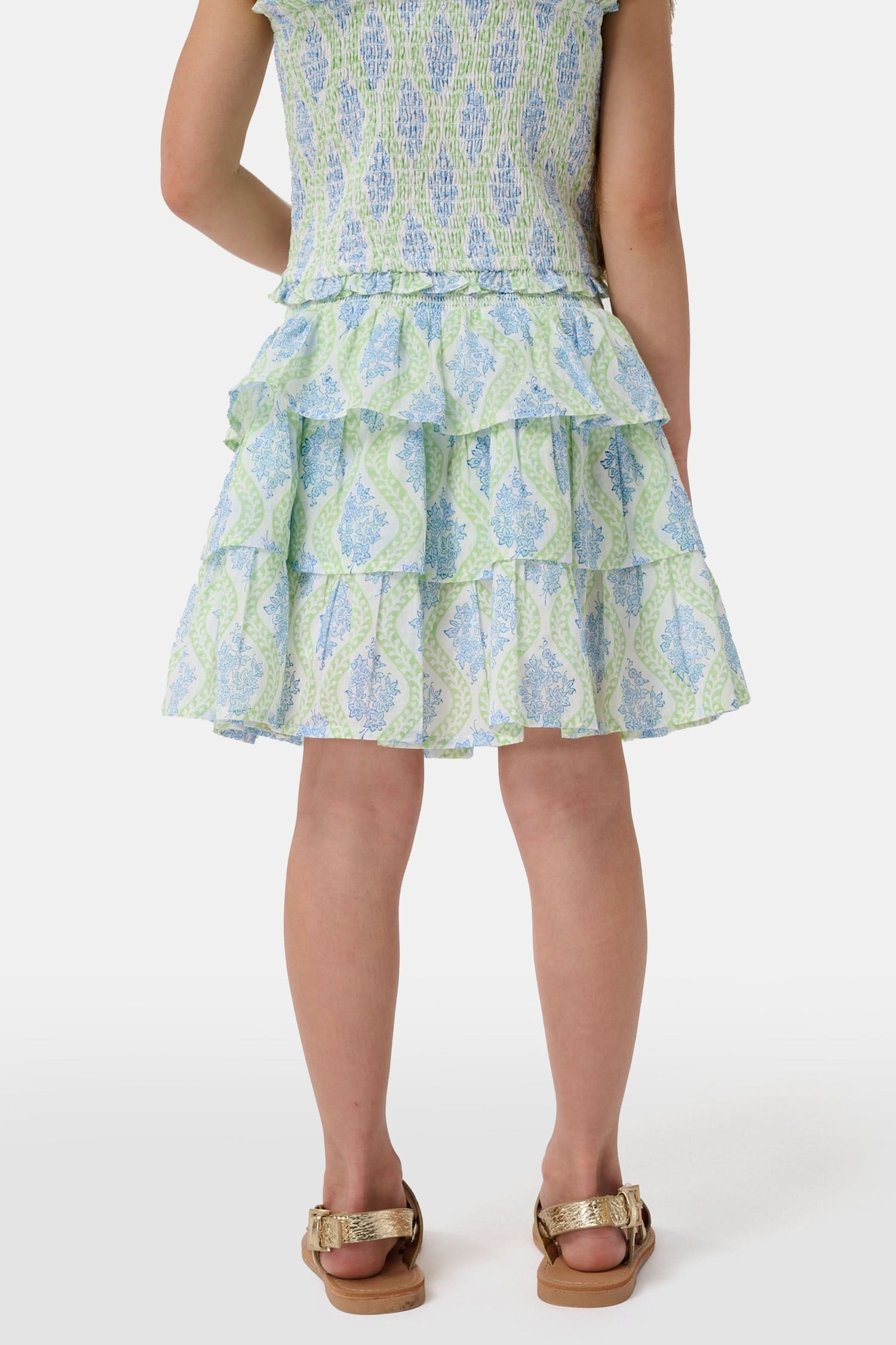 A smiling girl wears the Roller Rabbit Kids Zelda Floral Sami Skirt as she stands by a blue bicycle near a marina, surrounded by others in summer outfits with boats, water, and houses in the background under a clear sky.