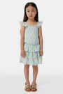 A smiling girl wears the Roller Rabbit Kids Zelda Floral Sami Skirt as she stands by a blue bicycle near a marina, surrounded by others in summer outfits with boats, water, and houses in the background under a clear sky.