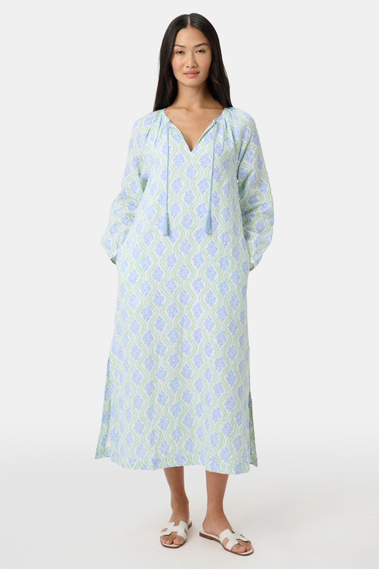 A woman faces forward in the Roller Rabbit Zelda Floral Shiv Caftan, a loose, ankle-length dress with a blue and green geometric print. She has long dark hair, white sandals, and stands against a plain white background.