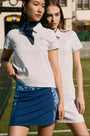 Two women stand on a tennis court wearing Roller Rabbit Pique Polo Shirts; one pairs hers with a blue patterned skirt and navy scarf, the other with a white skirt. A building appears in the background.