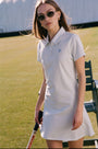 A woman in Roller Rabbit’s Pique Polo Dress and sunglasses stands on a tennis court holding a racket, with a bench and large white tent structure in the background.