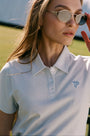 A woman in Roller Rabbit’s Pique Polo Dress and sunglasses stands on a tennis court holding a racket, with a bench and large white tent structure in the background.